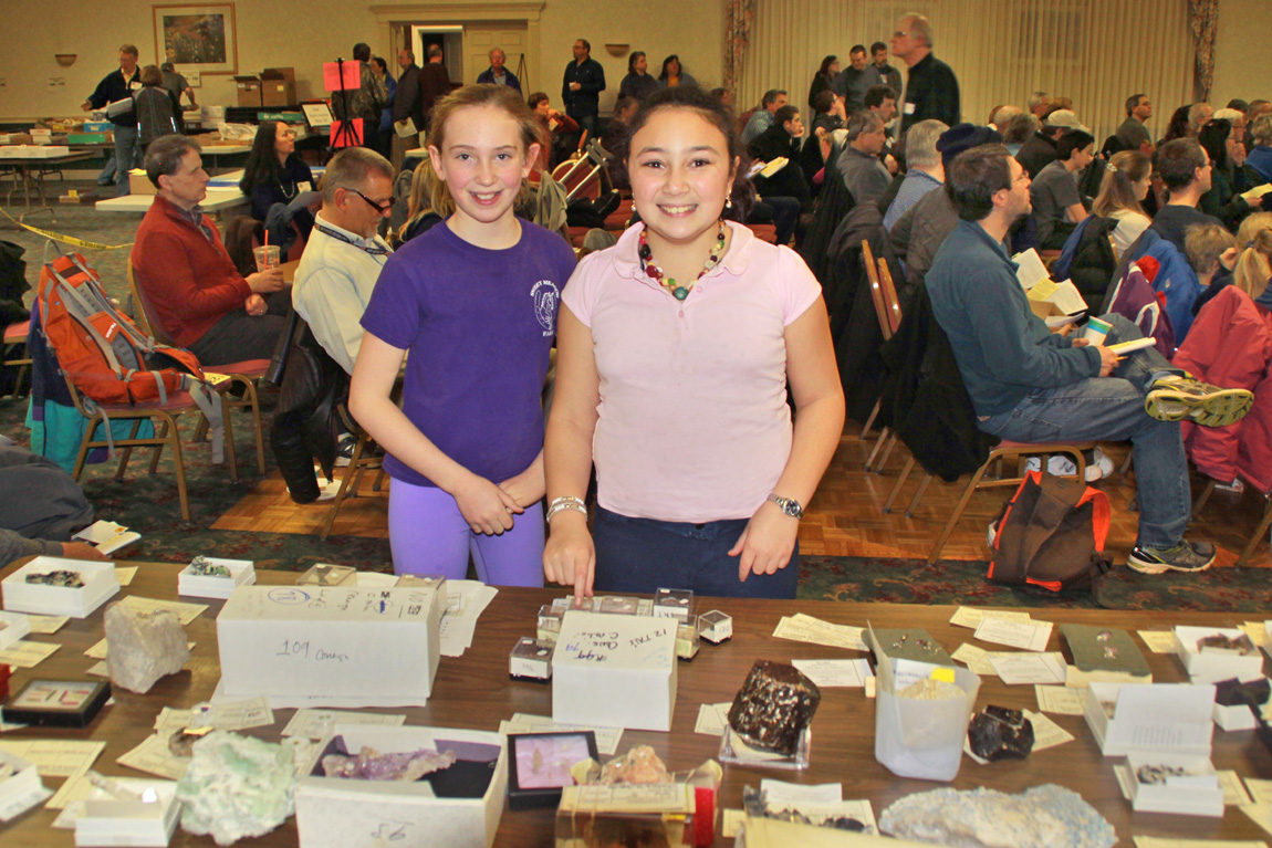 Two young girls with a table full of mineral specimens in front of them and an audience of seated bidders behind them